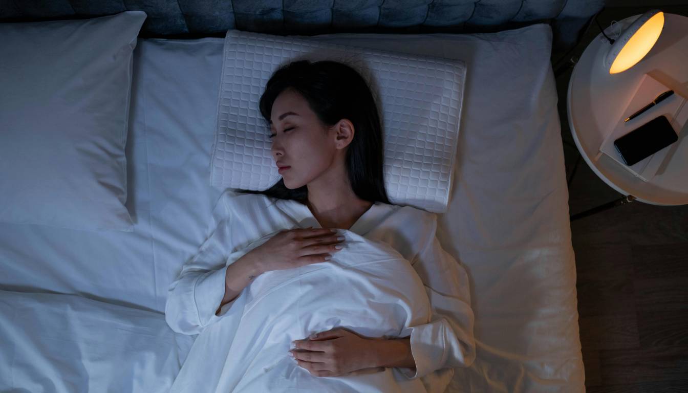 A person lying in bed with hands on the chest, covered with a white blanket, next to a nightstand with a lamp, a pen, and a smartphone.