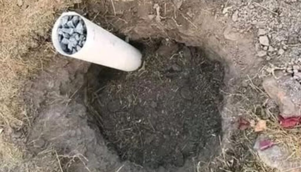 A white PVC pipe filled with gravel is inserted vertically into a hole dug in the ground.