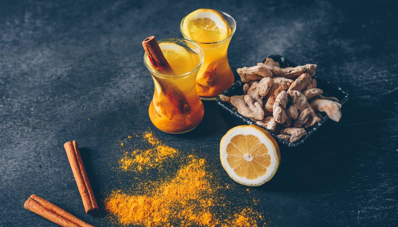 Two glasses of yellow-orange beverage with cinnamon sticks and lemon slices, surrounded by turmeric powder, a halved lemon, ginger roots, and cinnamon sticks on a dark surface.