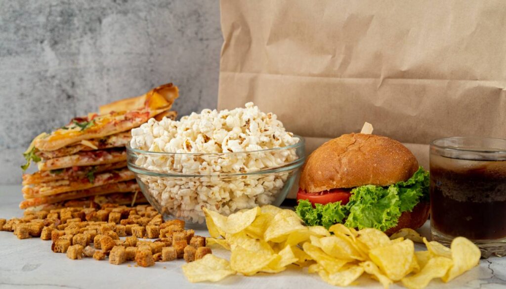 A variety of junk foods including pizza slices, popcorn, croutons, potato chips, a burger with lettuce and tomato, and a glass of soda are displayed on a table.