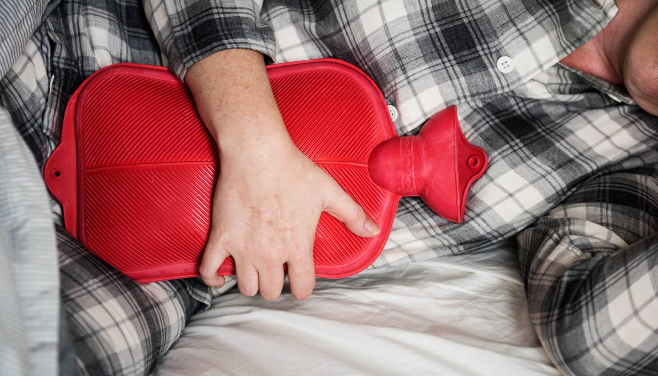 A person in plaid pajamas lying in bed, holding a red hot water bottle against their stomach.