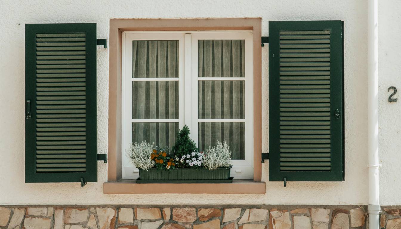 A window with green shutters, a flower box filled with various plants and flowers, and a stone wall beneath it