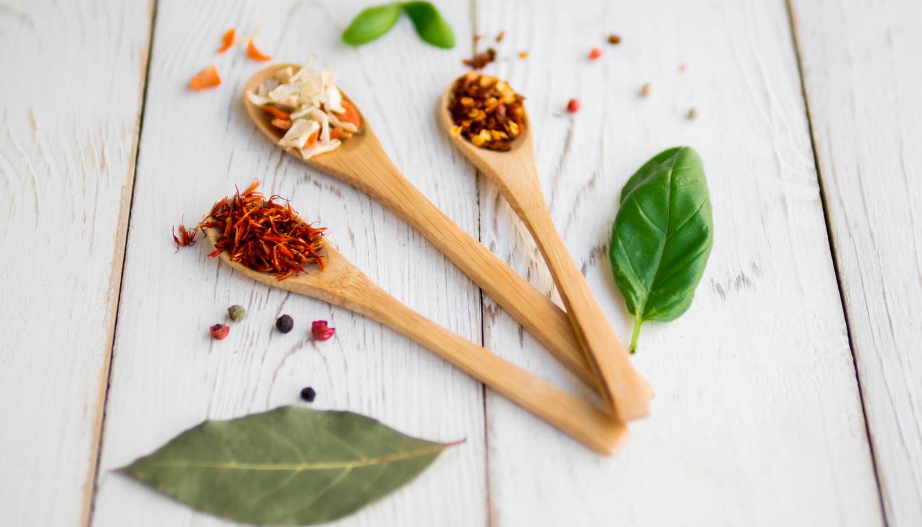 Three wooden spoons filled with different dried herbs and spices, surrounded by fresh basil and bay leaves on a white wooden surface.