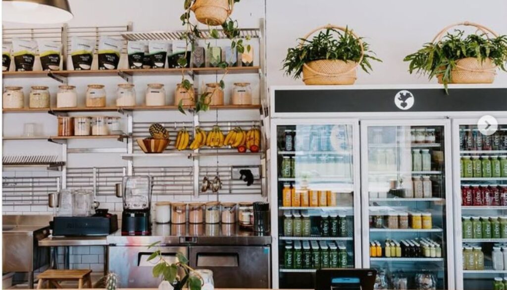 A cozy and modern herbal store with wooden shelves filled with various herbal products, a small round table with chairs, hanging plants, and a counter area with a neon sign that reads "kombucha bar" against a green wall.