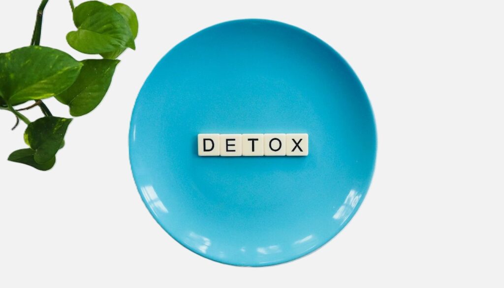 The word "DETOX" spelled out with letter tiles on a blue plate, with a green leafy plant visible on the side.