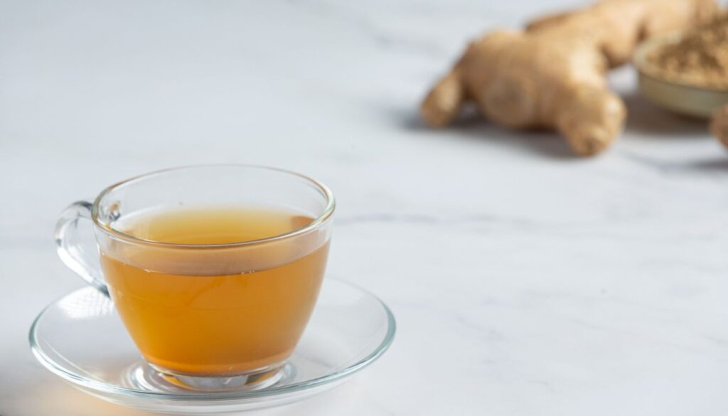 A glass cup of ginger tea placed on a saucer, surrounded by fresh ginger roots and ground ginger powder on a marble surface.