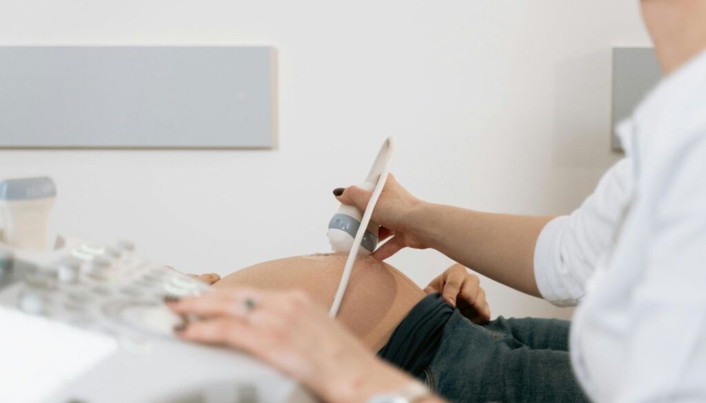 A pregnant woman sitting on a sofa, holding her belly and looking thoughtful, with a glass of water and prenatal vitamins on a table nearby.