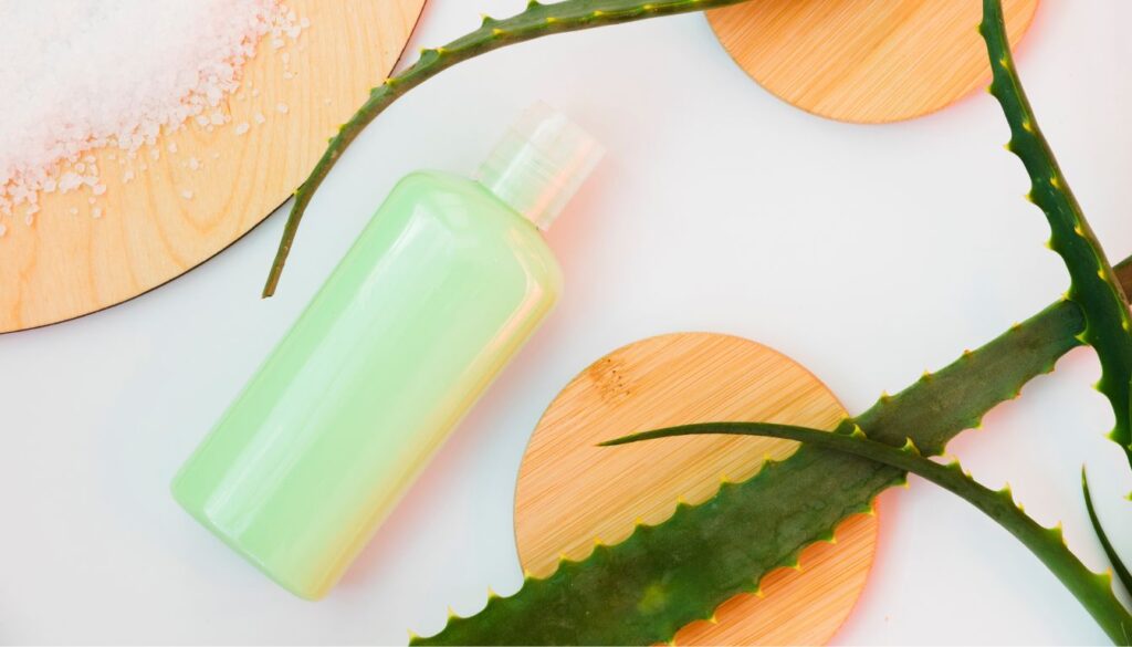 A green bottle of aloe vera gel on a white surface, surrounded by aloe vera leaves and coarse salt on a wooden coaster.