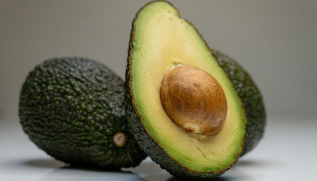 A halved avocado with its seed visible, placed beside a whole avocado on a wooden cutting board, surrounded by skincare products and a hairbrush.