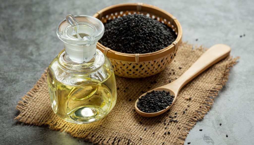 A glass bottle filled with dark, amber black cumin oil rests on burlap, accompanied by a wooden spoon and a woven basket of black cumin seeds.