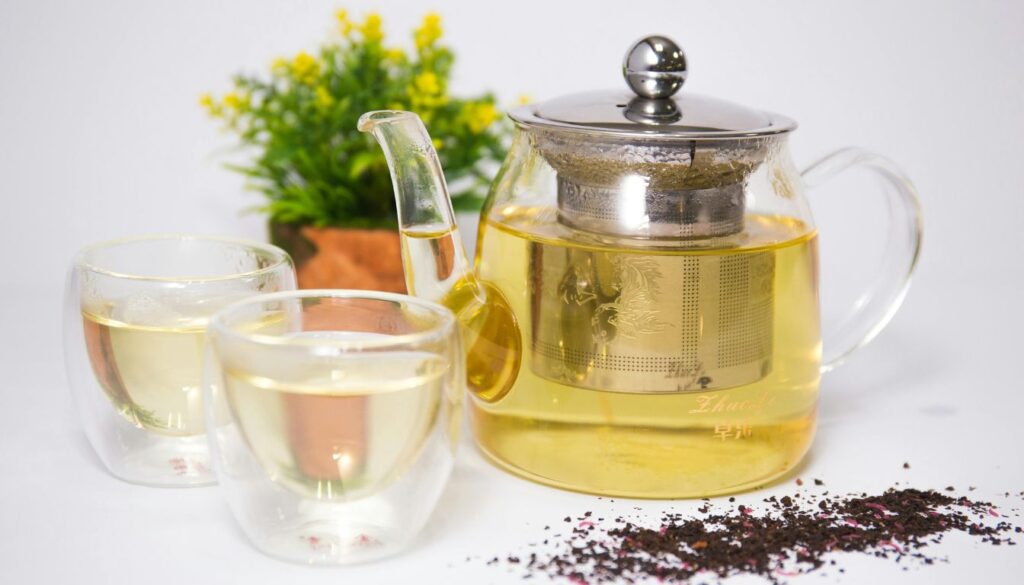 A steaming teacup and vintage teapot on a rustic table with scattered loose tea leaves.