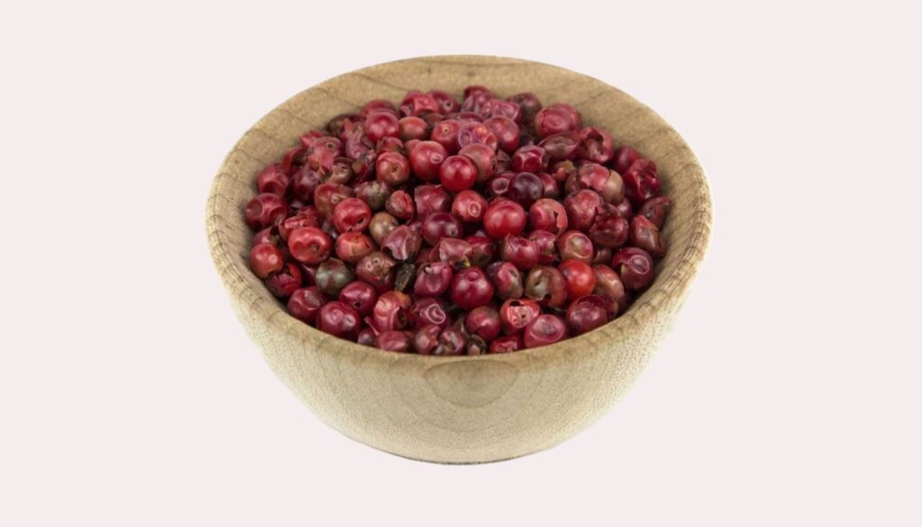 A wooden spoon filled with vibrant pink peppercorns resting on a rustic wooden table surrounded by scattered peppercorns.