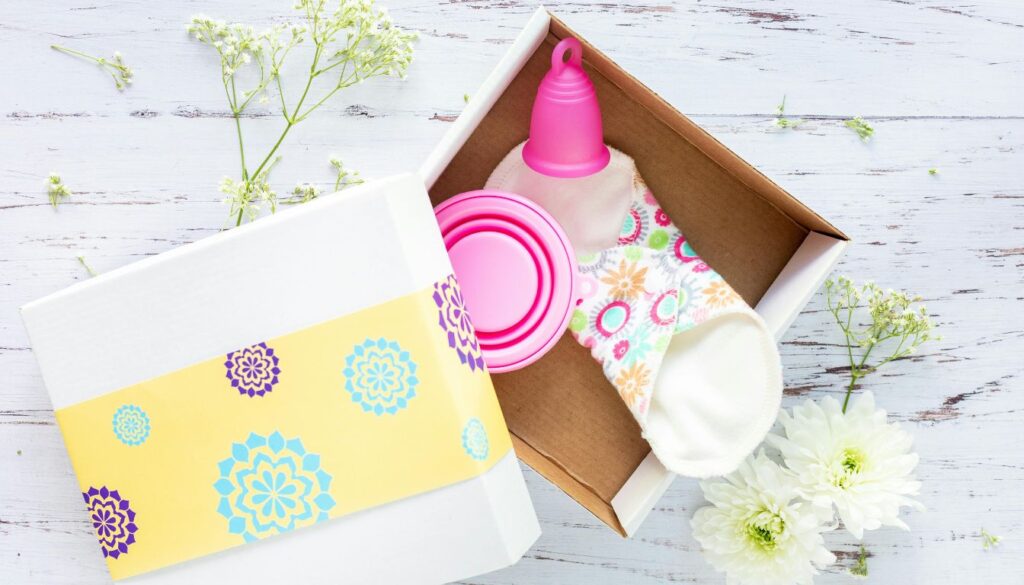 A gift box containing a pink menstrual cup, a collapsible pink cup, and a colorful patterned cloth pad, surrounded by white flowers and delicate greenery on a white wooden surface.