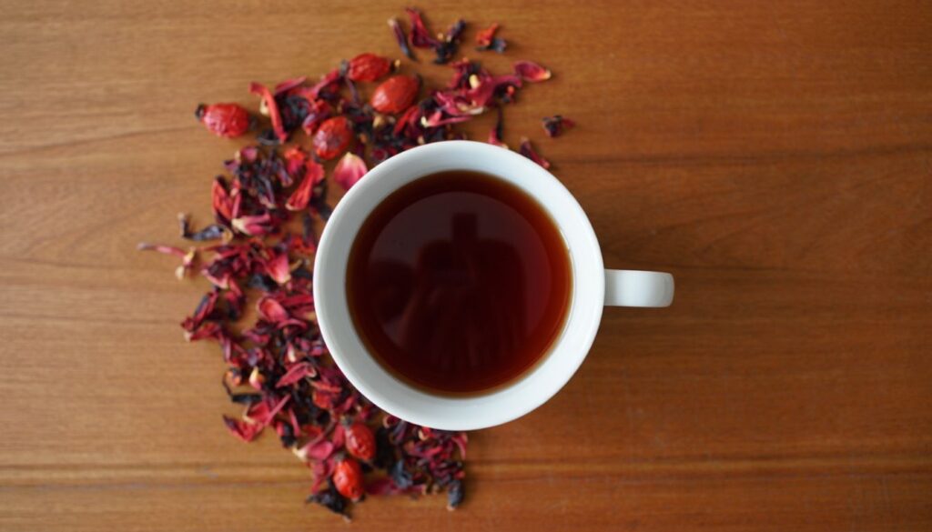 A white cup filled with hibiscus and rosehip tea on a wooden surface, surrounded by dried hibiscus petals and rosehips, showcasing the tea's vibrant red color.