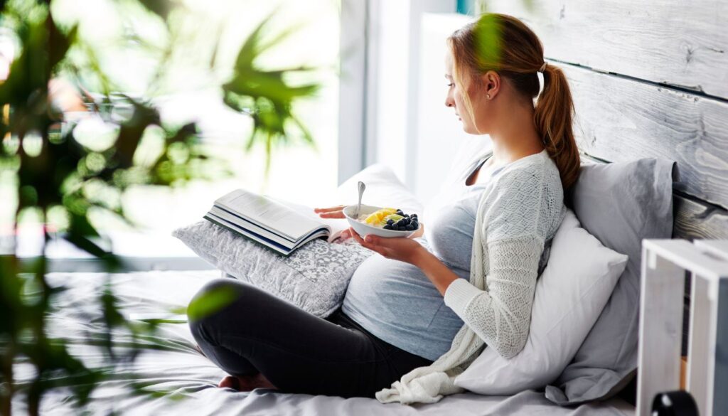 A pregnant woman seated comfortably on a couch, sipping water with a bowl of fruits on a nearby table.
