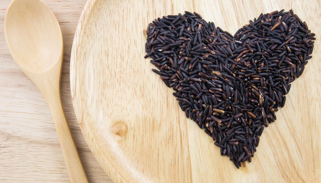A bowl of freshly cooked purple rice, garnished with herbs and placed on a wooden table alongside vegetables and a serving spoon.
