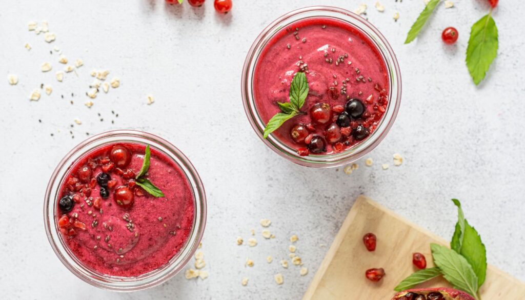 Two glasses of vibrant low-carb smoothie garnished with fresh berries, mint leaves, and chia seeds, placed on a light surface surrounded by fruits.