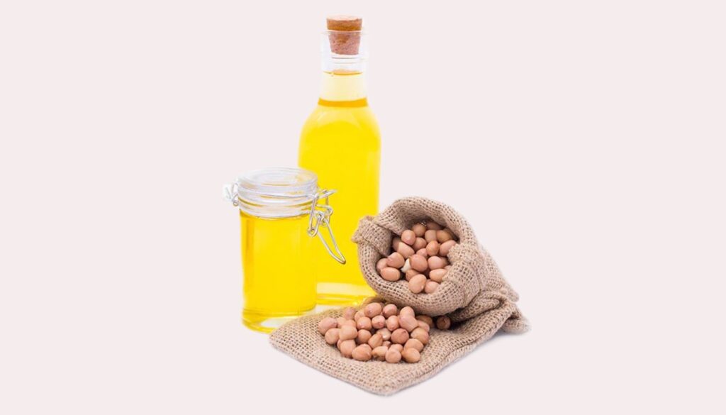 A bottle of groundnut oil accompanied by scattered shelled and unshelled peanuts on a wooden surface.