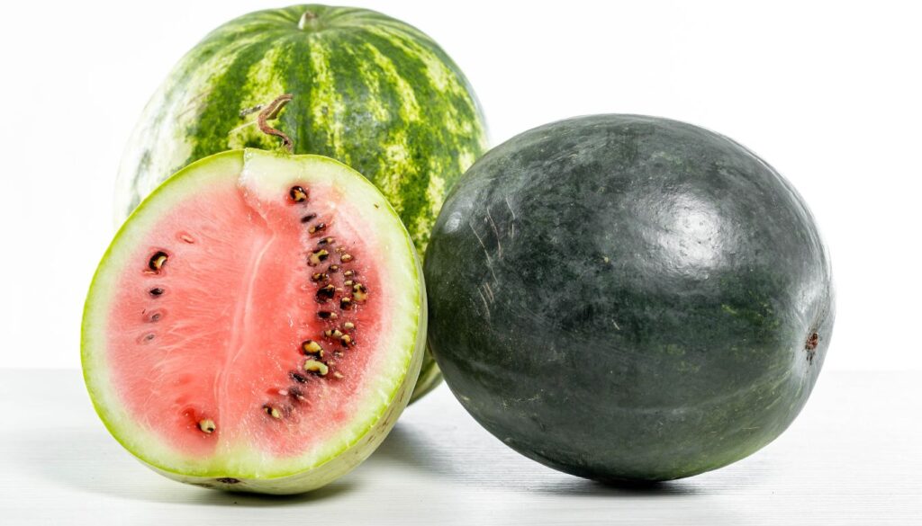 Freshly sliced watermelon pieces arranged on a wooden platter alongside whole watermelons, set on a picnic table under a sunny sky.