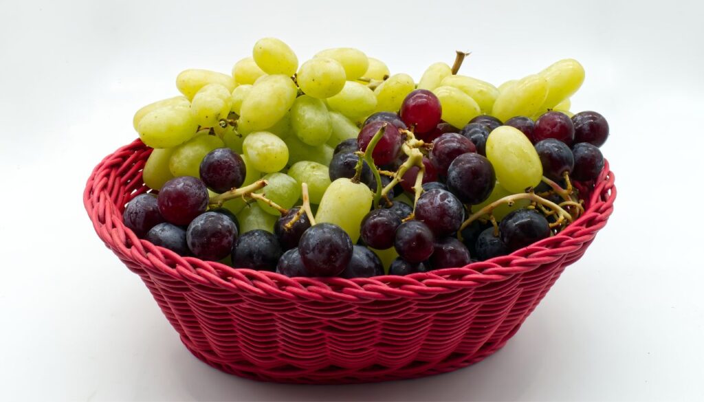 A wooden bowl filled with vibrant red grapes, surrounded by more grapes scattered on a white wooden surface.