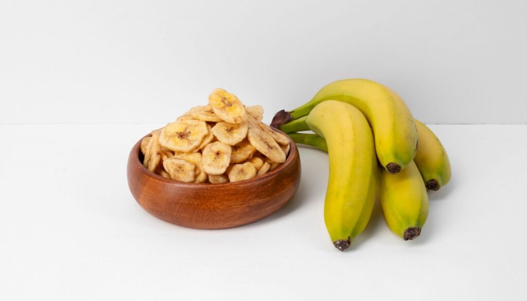 A bowl filled with crispy banana chips, golden in color, placed on a wooden surface with a few scattered chips around it.