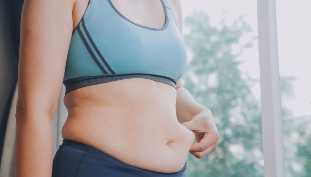 A woman in athletic gear stands in front of a window, preparing for a workout, with her midsection visible, highlighting tummy fat.
