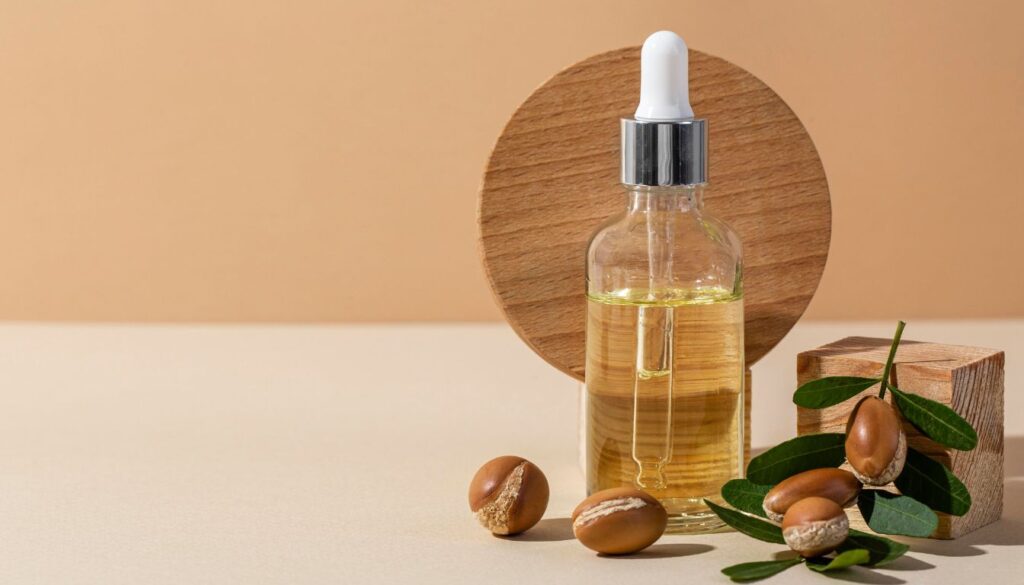 A small dropper bottle filled with golden argan oil, placed next to scattered argan nuts and a wooden decorative cube on a neutral surface.