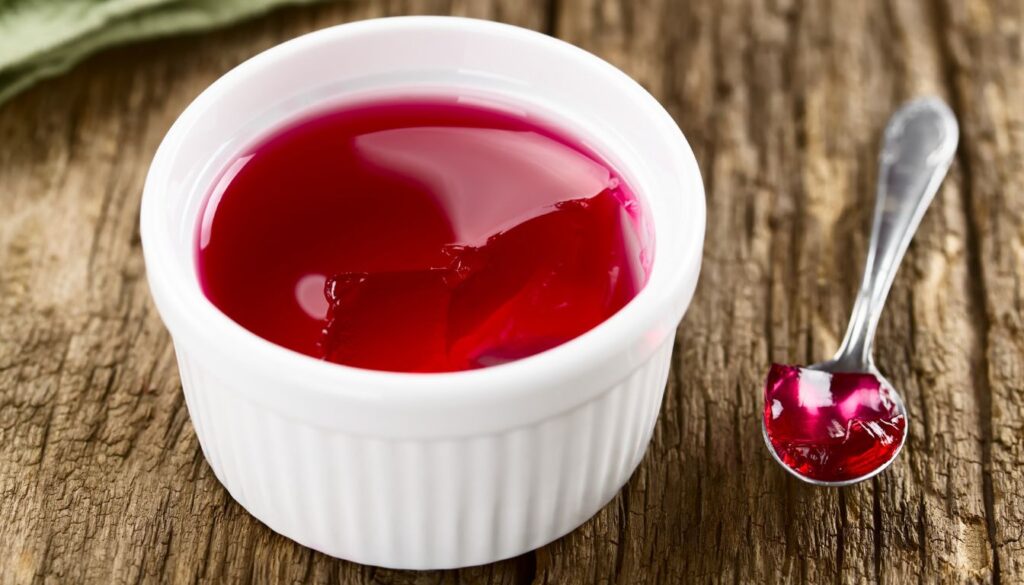 A white ramekin filled with red gelatine sits on a wooden surface. A spoon with a portion of the red gelatine is placed next to the ramekin.
