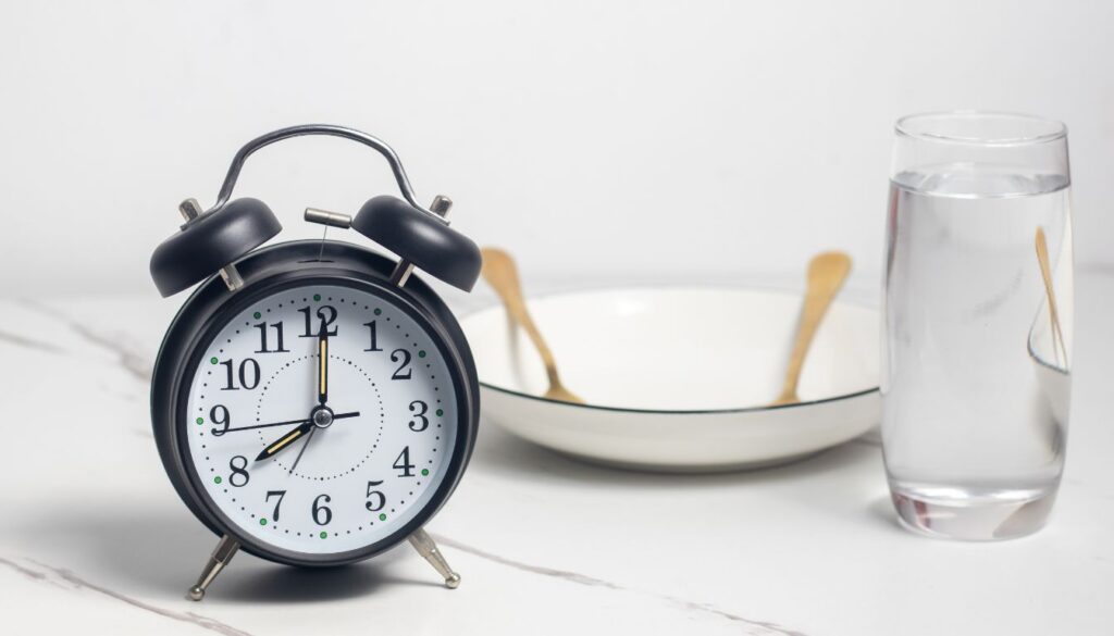 A black analog clock showing 10:10 sits on a marble countertop. Behind it, an empty white bowl with two wooden spoons is placed next to a clear glass filled with water.
