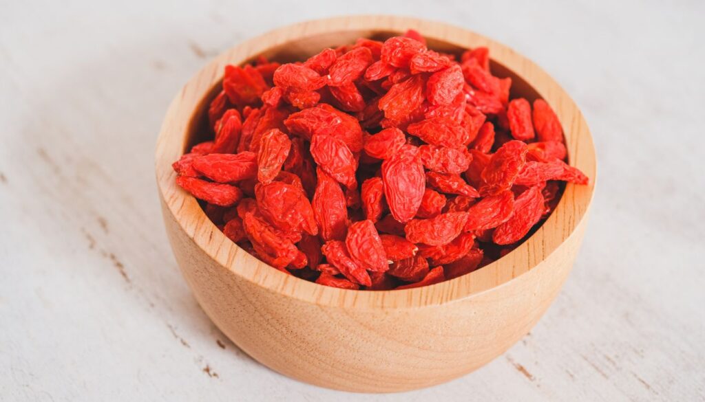 A wooden bowl filled with vibrant red goji berries, with additional berries scattered around the bowl on a light wooden surface.