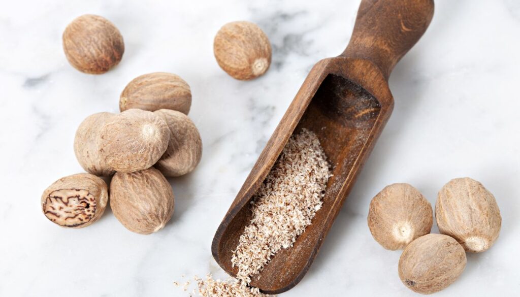 A close-up of whole and ground nutmeg on a rustic wooden surface, with a nutmeg grater beside them, highlighting the spice’s rich texture and earthy tones.