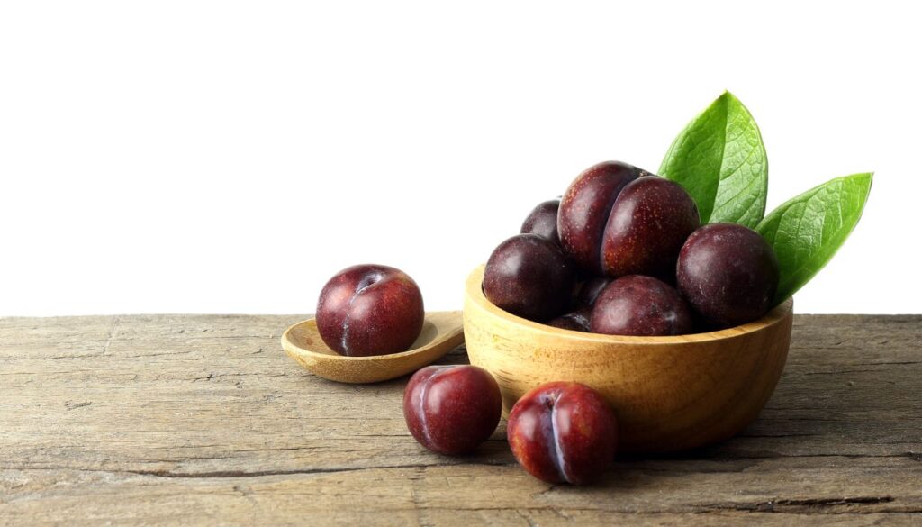 A wooden bowl filled with fresh, red plums, with additional plums scattered around the bowl on a white wooden surface.