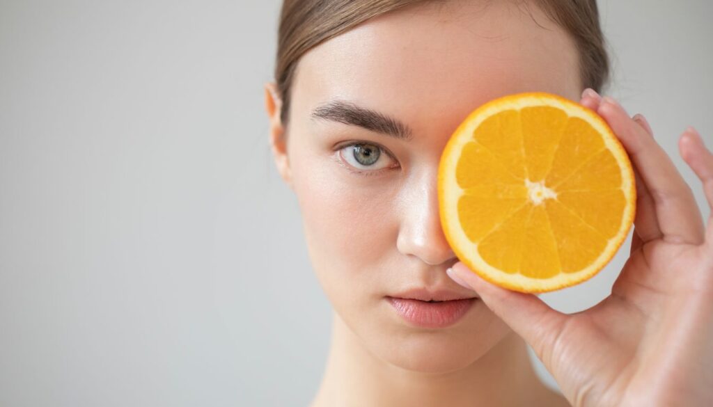 A woman holds an orange slice over her left eye while looking directly into the camera with her right eye, showcasing a playful and vibrant expression.