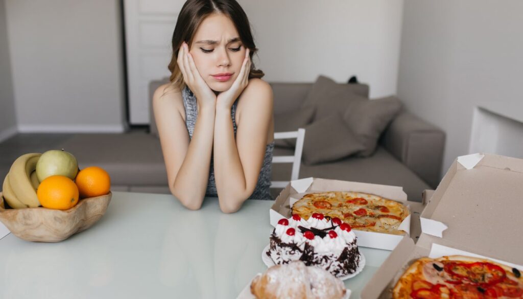 A reflective person sitting at a table with a spread of fatty foods (such as pizza, cake, and donuts), symbolizing an exploration of food cravings.