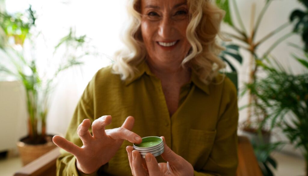 Person in a green shirt applying green herbal cream from a small container, surrounded by lush green plants.