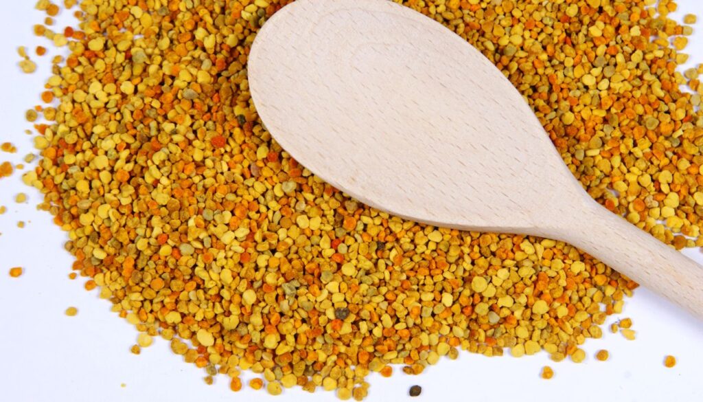 A wooden spoon resting on a pile of bee pollen granules, which are yellow, orange, and brown in color.