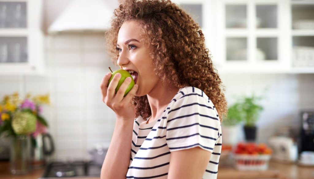 A woman in her kitchen enjoying a green apple, highlighting healthy eating habits and mindful nutrition.