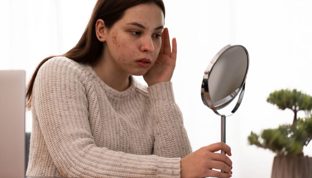 Person in beige sweater holding a round mirror and touching their face near a laptop and potted plant.