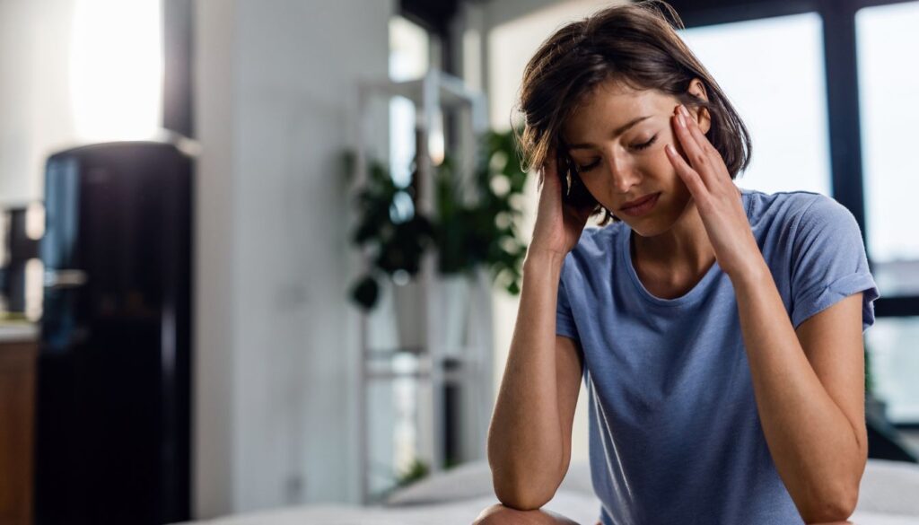 Person in a blue shirt holding their head, seated indoors with a blurred background of plants and large windows.