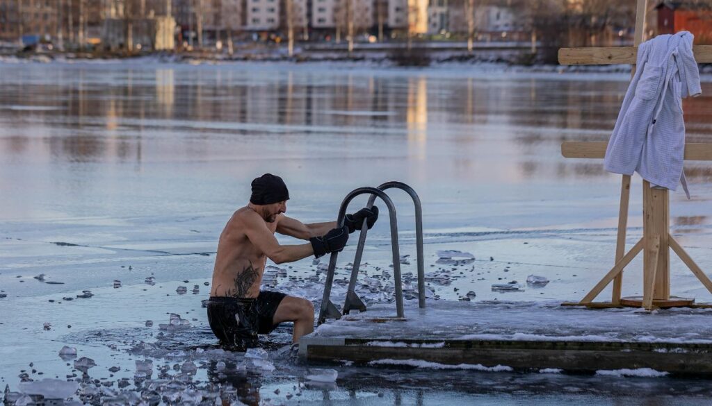 A man climbs a ladder to exit a frozen lake, surrounded by icy water and winter scenery.