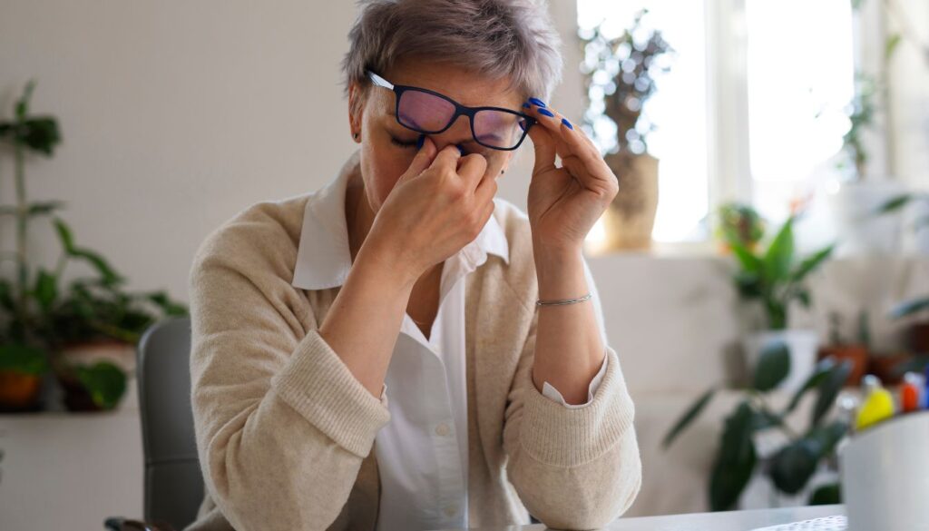 Person in a beige cardigan holding glasses at a desk, surrounded by green plants and soft natural light.