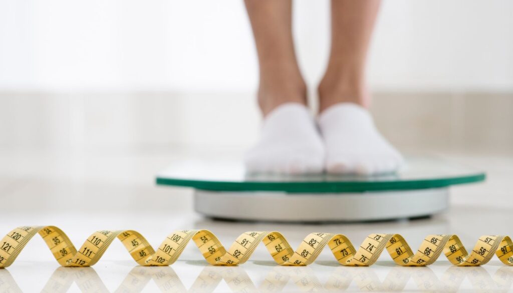 A person standing on a weighing scale with a measuring tape placed in front, symbolizing weight management and fitness goals.