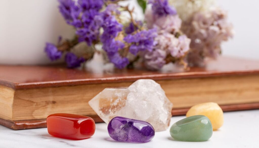 A collection of colorful crystals and herbs arranged on a wooden book. The crystals include red, purple, green, and yellow stones, along with a larger clear quartz crystal. Behind the crystals, there are vibrant purple and white flowers.