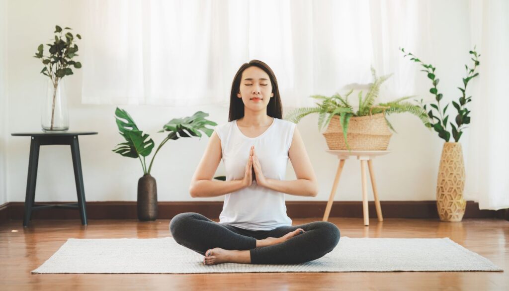 A person sitting cross-legged on a yoga mat in a serene room, surrounded by lush green plants, practicing yoga with hands in a prayer position.