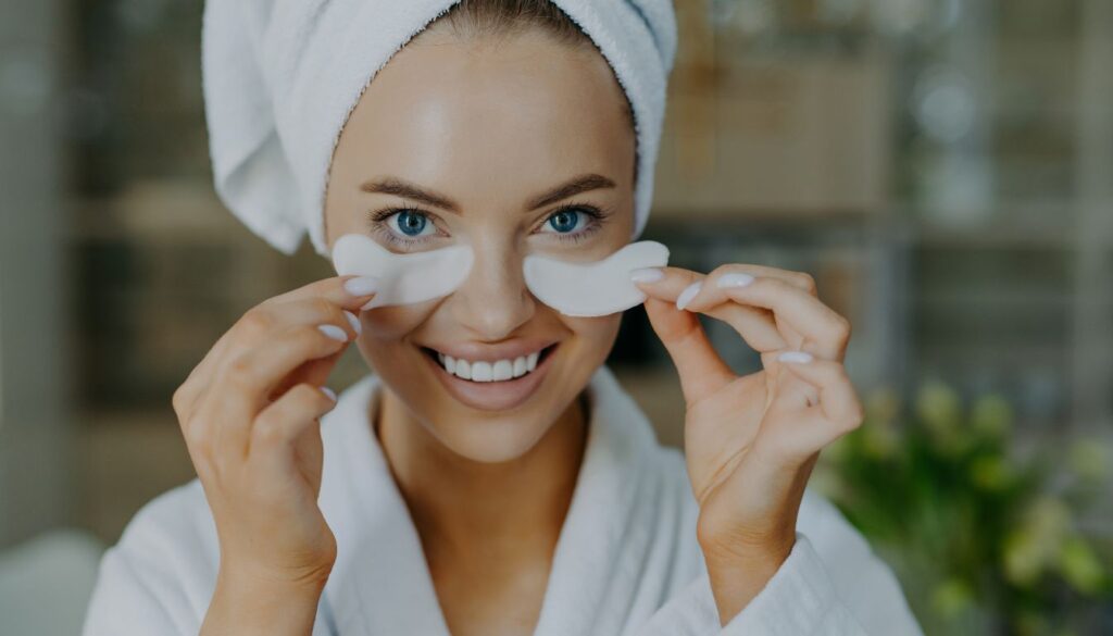 A person wearing a white bathrobe and a towel on the head is holding two cotton pads near the eyes.