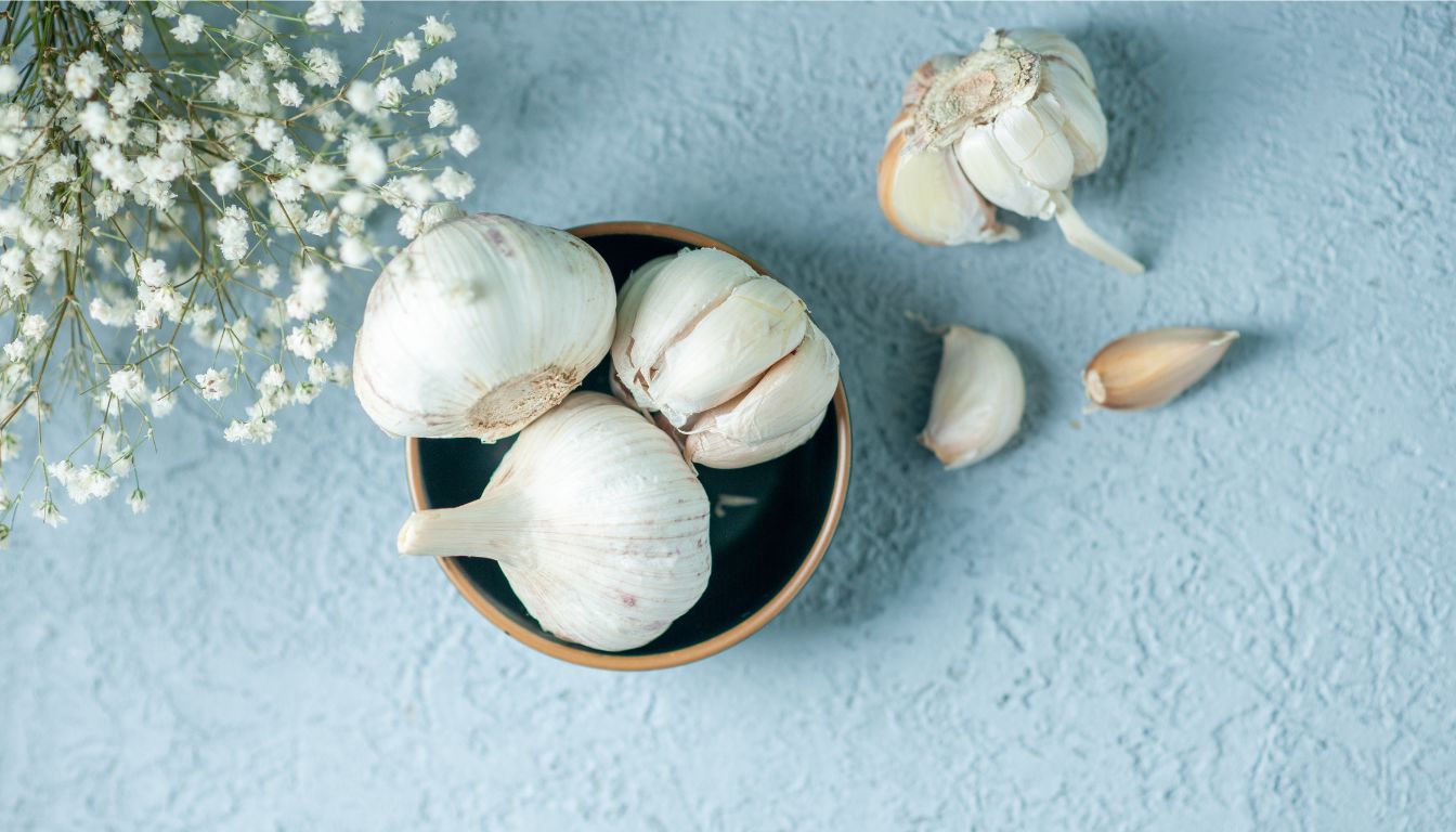 Three whole garlic bulbs in a ceramic bowl with loose cloves and baby's breath flowers on a textured blue surface.