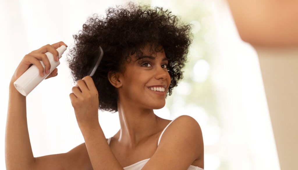 Person with curly hair using a spray bottle and comb during a hair care routine, wearing a white sleeveless top in a softly lit indoor setting.