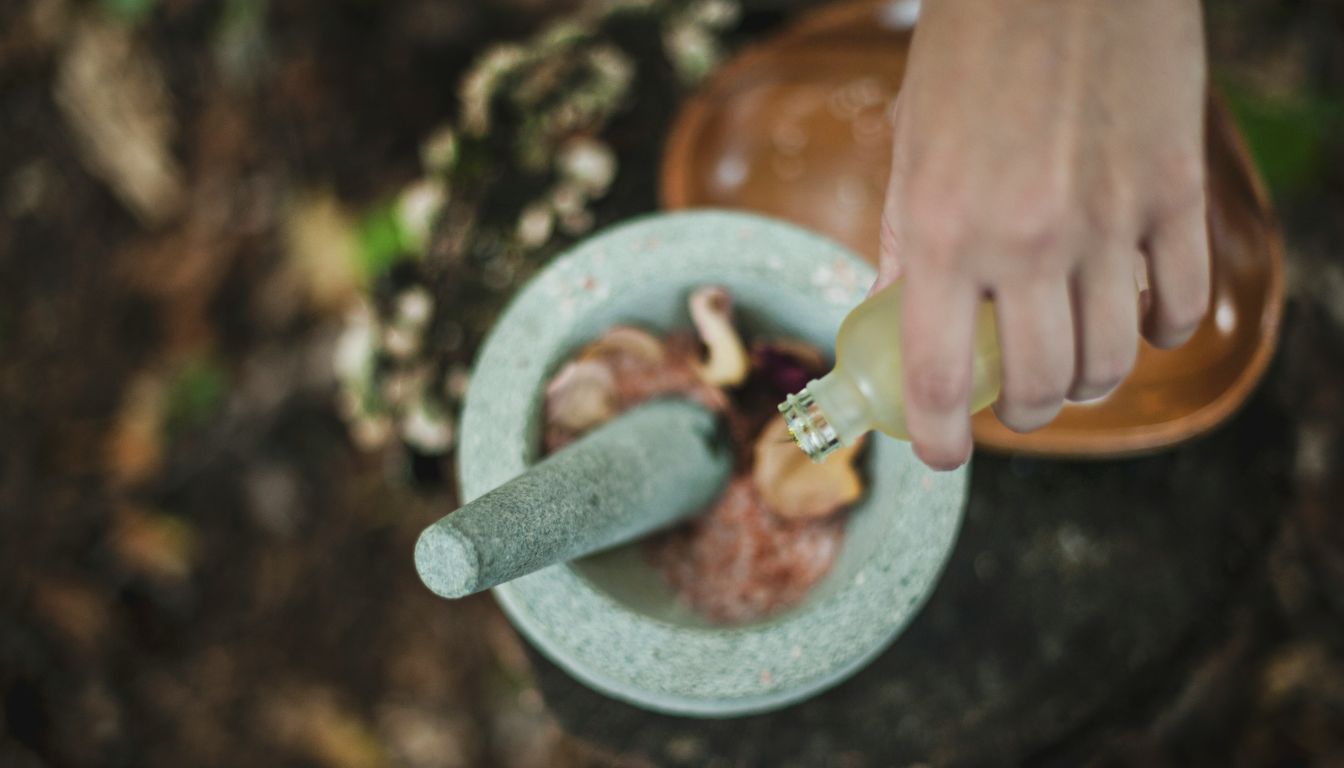 Hand pouring liquid into a mortar filled with dried herbs outdoors, representing traditional herbal preparation.