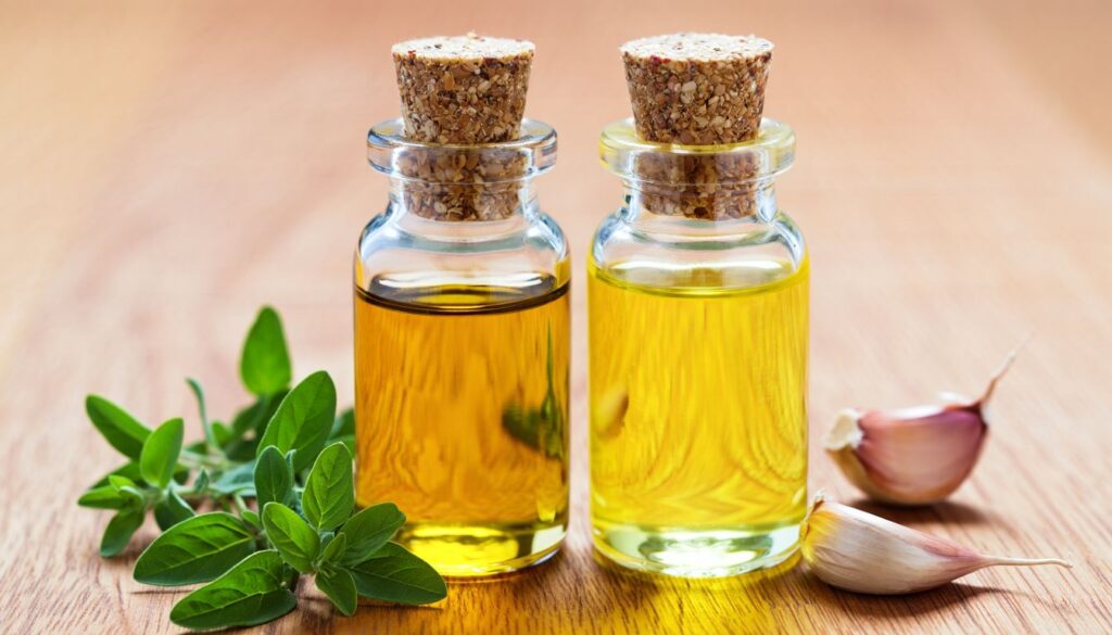 Two cork-stopped glass bottles of pale yellow oil on a wooden surface, one beside fresh oregano leaves and the other beside garlic cloves.